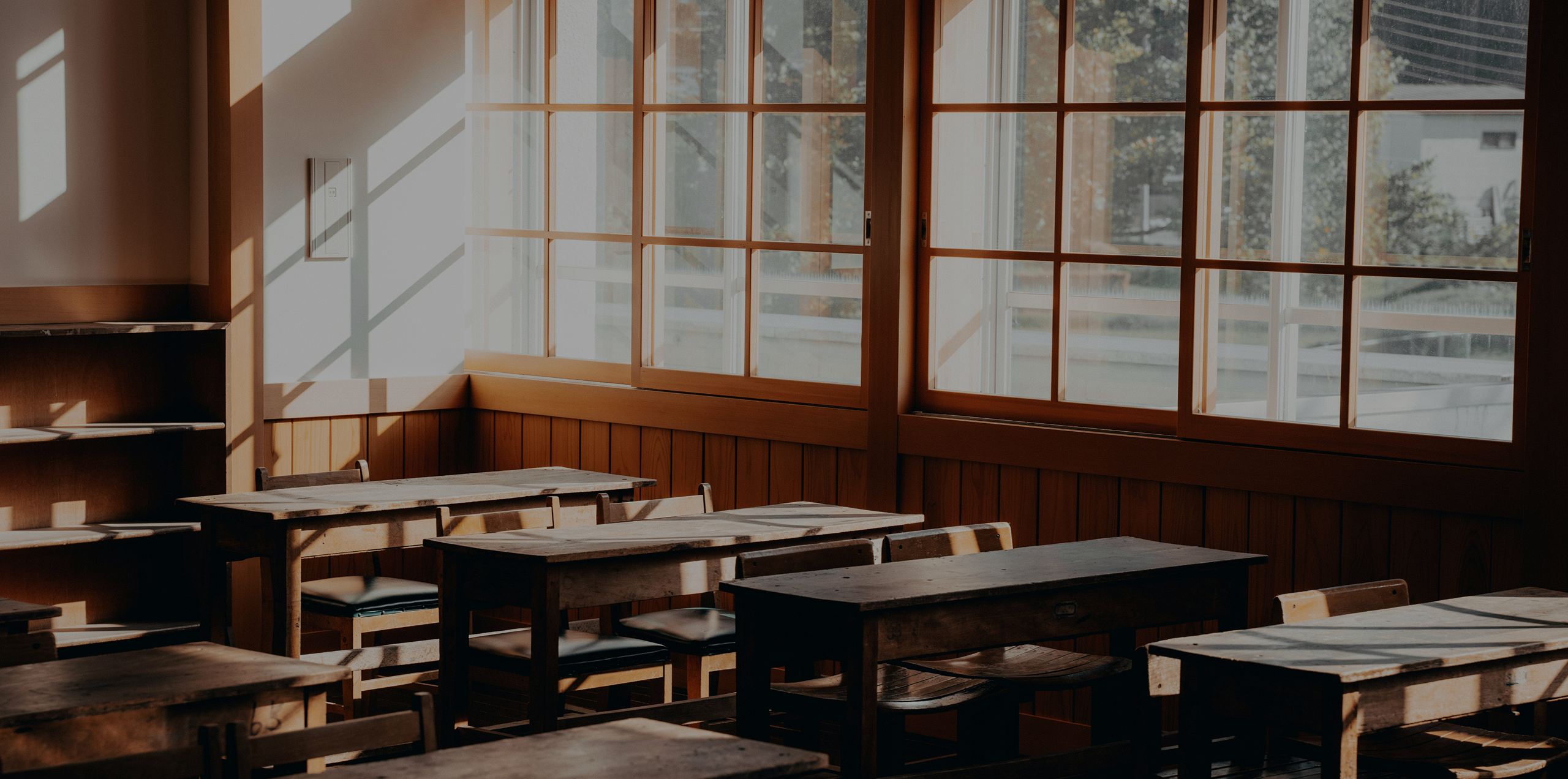 Photo of a elementary school classroom with empty wooden desks and sun shining through the windows.